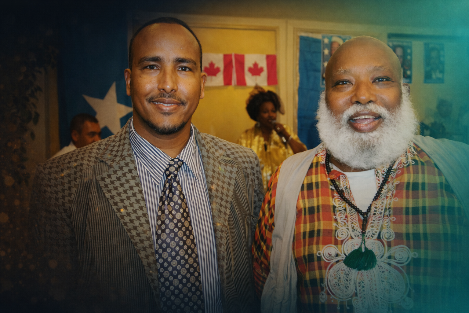 Two men smiling, one wearing a patterned suit and tie, the other with a white beard and colorful traditional attire, with flags in the background.