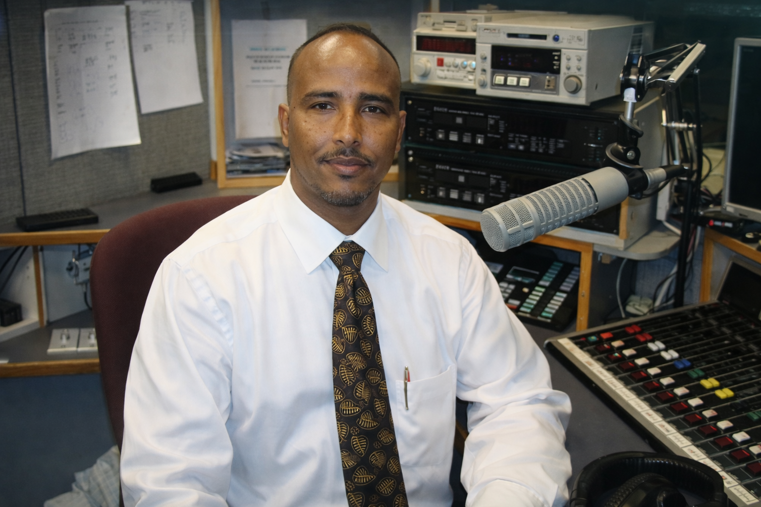 Man wearing white shirt and patterned tie sitting in a radio studio with microphone and audio mixing board.